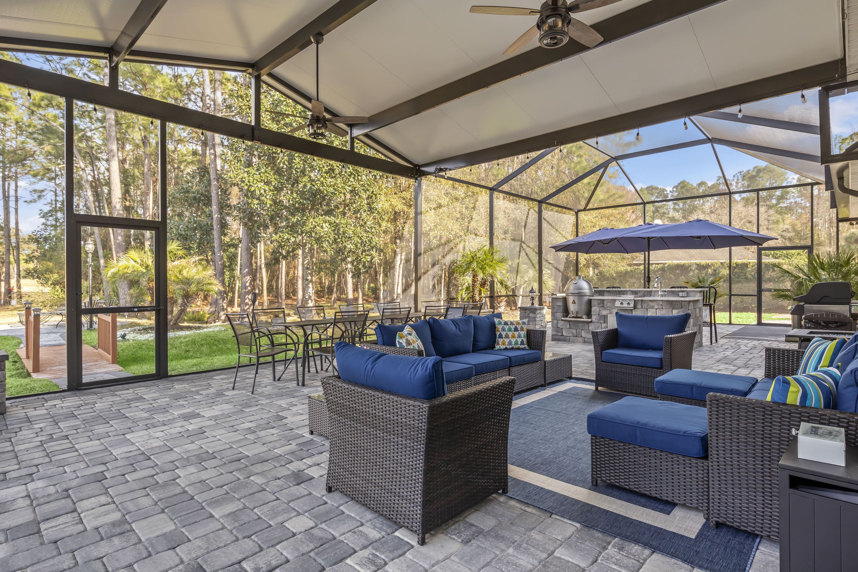 Spacious and stylish screened outdoor living space featuring wicker furniture with blue cushions, dining and lounge areas, ceiling fans, and a built-in grill station - Impact Enclosures - Outdoor Enclosures Jacksonville